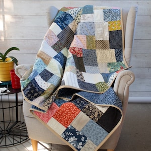 May include: A patchwork quilt featuring a mix of blue, green, red and white fabrics. The quilt is folded and draped over a light grey armchair.