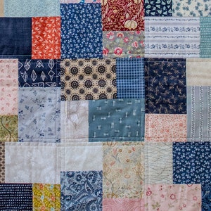 May include: Close-up of a patchwork quilt with a variety of patterned fabrics. The quilt features squares of denim, floral prints, and geometric designs in shades of blue, red, and beige. The quilt is stitched together with white thread.