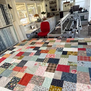 May include: A colourful patchwork quilt in progress, with a mix of patterned fabrics in shades of red, blue, and floral designs. The quilt is spread out on a large white surface in a bright room, with fabric bolts on shelves in the background.