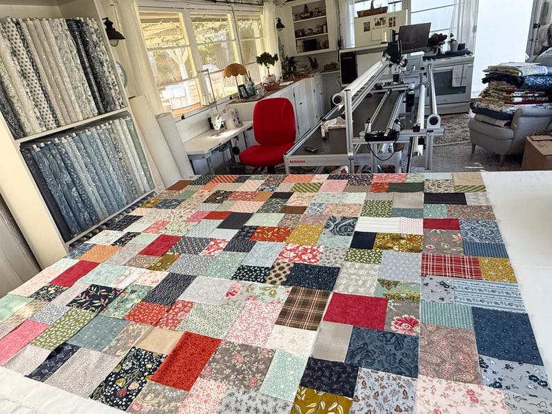 May include: A colourful patchwork quilt in progress, with a mix of patterned fabrics in shades of red, blue, and floral designs. The quilt is spread out on a large white surface in a bright room, with fabric bolts on shelves in the background.
