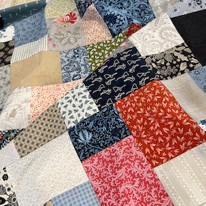 May include: A patchwork quilt featuring a mix of patterned fabrics in blue, red, beige, and black. The quilt showcases floral, geometric, and abstract designs, creating a visually diverse and textured surface. The quilt is made of multiple fabric squares.