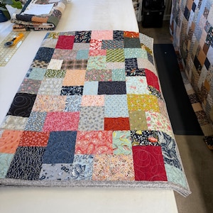 May include: A patchwork quilt featuring a mix of patterned fabric squares in red, blue, pink, and green. The quilt is displayed on a white surface, highlighting the detailed design and stitching. The quilt measures approximately 152 cm long and 122 cm wide.