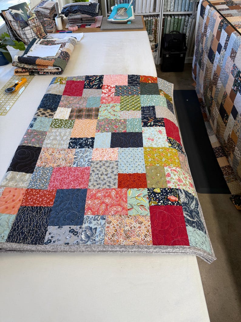 May include: A patchwork quilt featuring a mix of patterned fabric squares in red, blue, pink, and green. The quilt is displayed on a white surface, highlighting the detailed design and stitching. The quilt measures approximately 152 cm long and 122 cm wide.