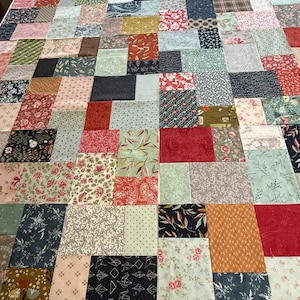 May include: A patchwork quilt in progress, with a variety of fabric squares in shades of red, blue, green, and cream. The quilt top is laid out on a white surface, showcasing floral, geometric, and patterned designs. The quilt is a mix of textures and colours.