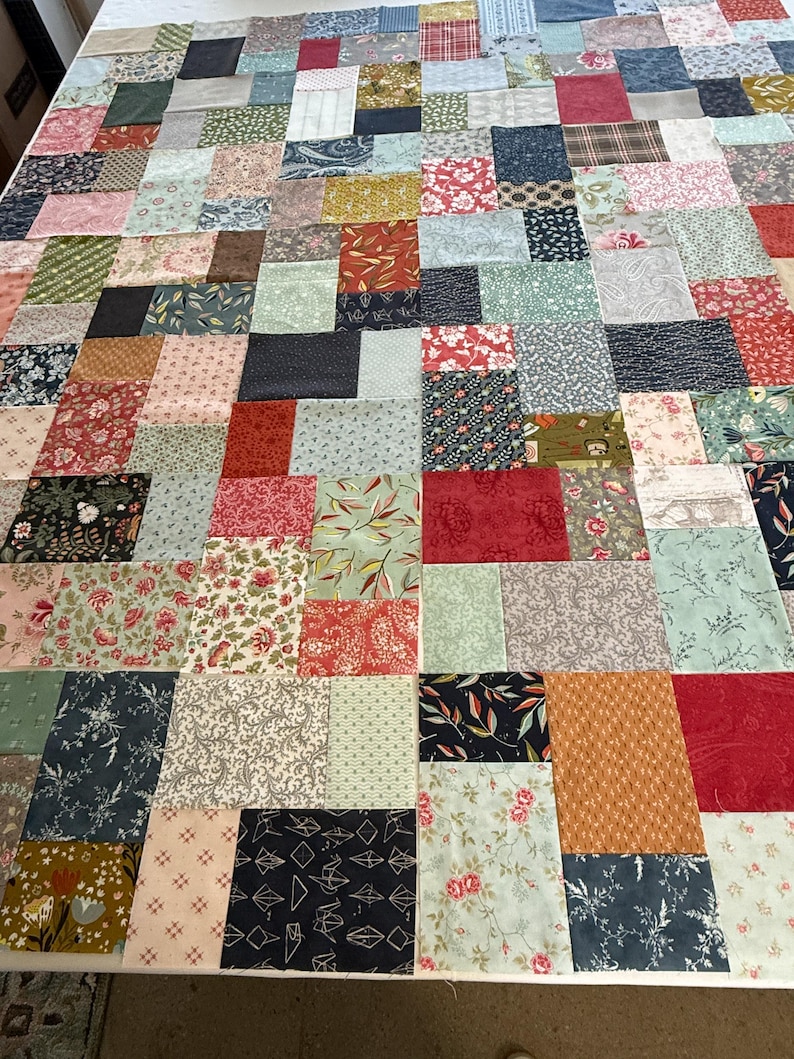 May include: A patchwork quilt in progress, with a variety of fabric squares in shades of red, blue, green, and cream. The quilt top is laid out on a white surface, showcasing floral, geometric, and patterned designs. The quilt is a mix of textures and colours.