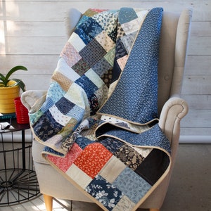 May include: A patchwork quilt in shades of blue, red, and beige, draped over a grey armchair. The quilt has a solid blue floral backing and a beige border. The quilt is made of various patterned fabrics. The armchair has wooden legs.