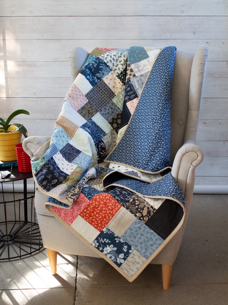 May include: A patchwork quilt in shades of blue, red, and beige, draped over a grey armchair. The quilt has a solid blue floral backing and a beige border. The quilt is made of various patterned fabrics. The armchair has wooden legs.
