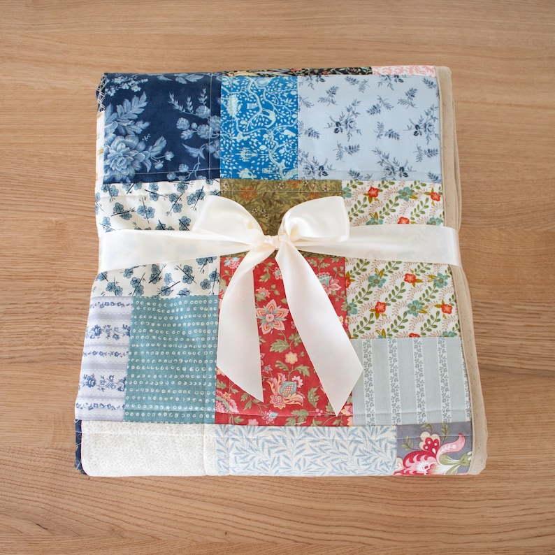 May include: A folded patchwork quilt tied with a cream ribbon bow. The quilt is made from various patterned fabrics in blue, red, and cream. The quilt is neatly folded, suggesting it is ready to be given as a gift.