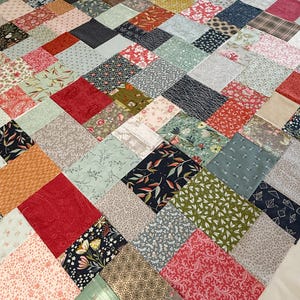 May include: A patchwork quilt in progress, with diamond-shaped fabric squares. The quilt top displays a mix of floral, geometric, and solid patterns in red, blue, green, and cream. The quilt is a handmade item.
