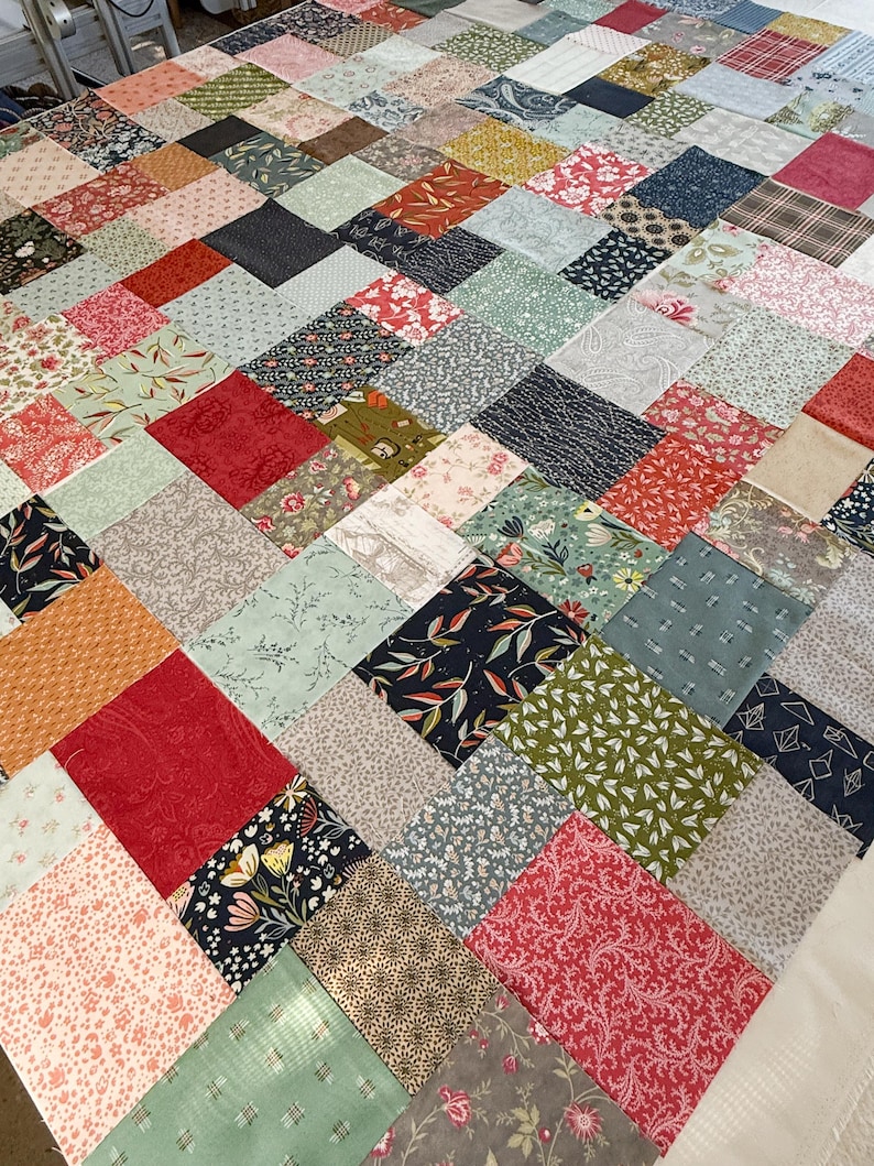May include: A patchwork quilt in progress, with diamond-shaped fabric squares. The quilt top displays a mix of floral, geometric, and solid patterns in red, blue, green, and cream. The quilt is a handmade item.