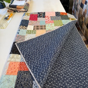 May include: A patchwork quilt featuring a mix of patterned fabric squares in pink, blue, green, and brown. The quilt is partially folded, showing a navy blue backing with a white paisley design. The edges are finished with a light grey binding.