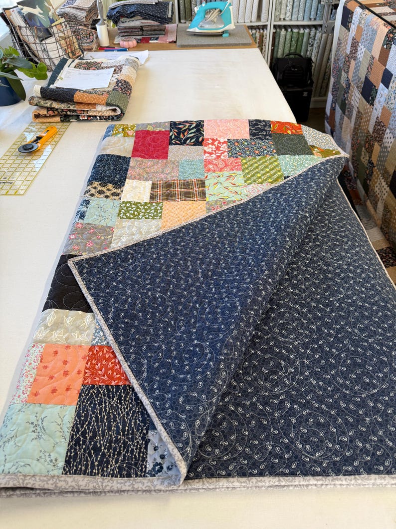 May include: A patchwork quilt featuring a mix of patterned fabric squares in pink, blue, green, and brown. The quilt is partially folded, showing a navy blue backing with a white paisley design. The edges are finished with a light grey binding.