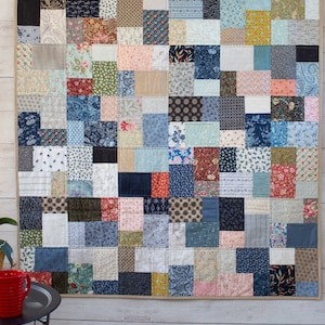 May include: A patchwork quilt featuring a mix of patterned fabric squares in blues, reds, beiges, and greens. The rectangular quilt is displayed against a white wall. It is held in place by two black clips.