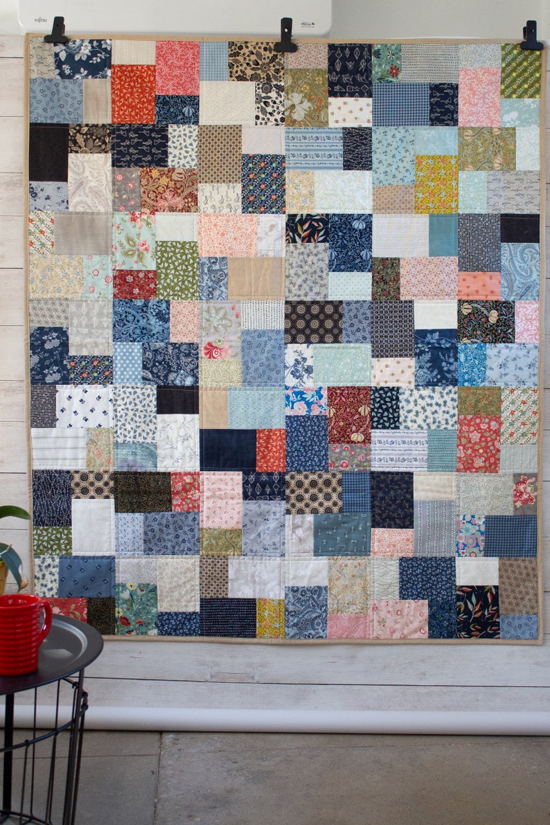 May include: A patchwork quilt featuring a mix of patterned fabric squares in blues, reds, beiges, and greens. The rectangular quilt is displayed against a white wall. It is held in place by two black clips.