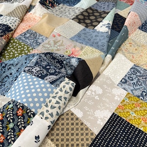 May include: A patchwork quilt featuring a variety of patterned fabrics in shades of blue, beige, and floral designs. The quilt is partially rolled up, showing the different fabric patterns. The quilt is a handmade item.