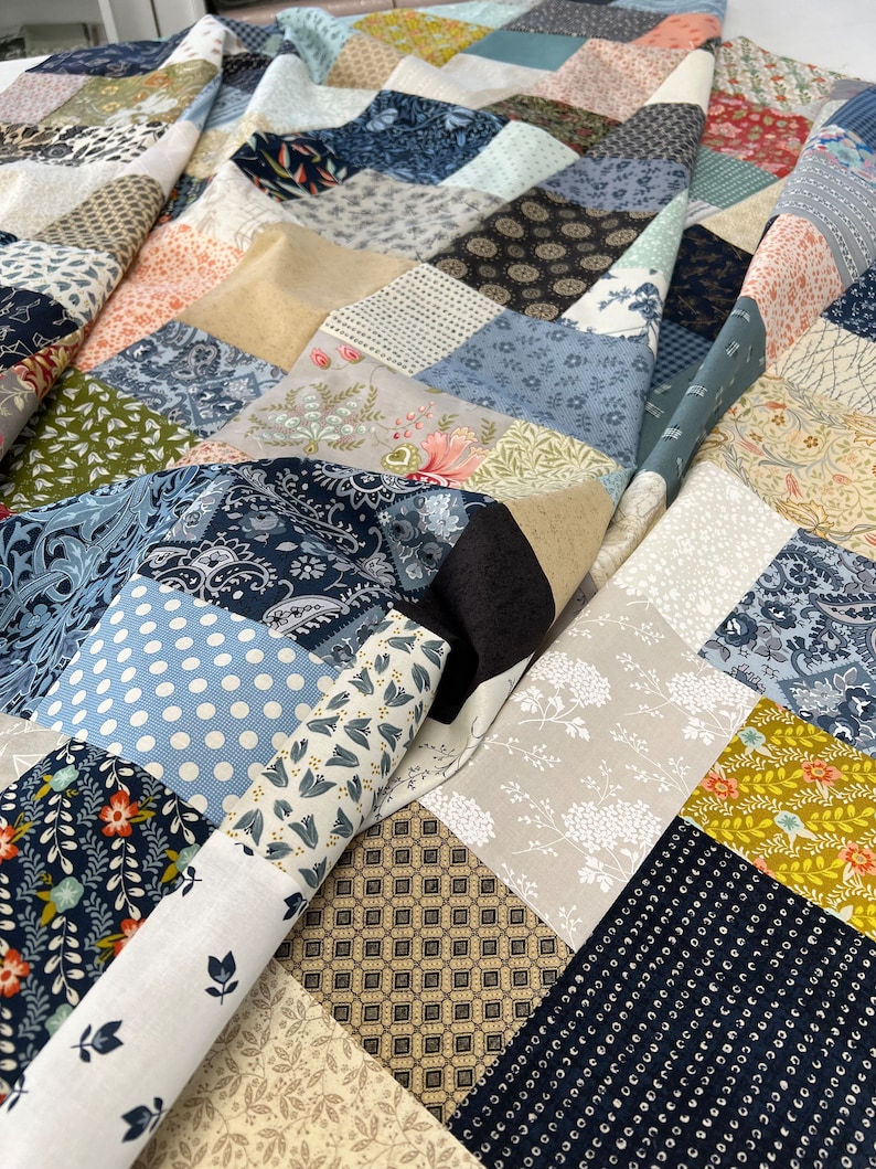 May include: A patchwork quilt featuring a variety of patterned fabrics in shades of blue, beige, and floral designs. The quilt is partially rolled up, showing the different fabric patterns. The quilt is a handmade item.