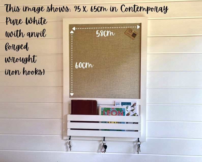 May include: White wall mounted message board with a cork surface and a shelf below. The board measures 23.6 inches by 25.6 inches. The shelf has two hooks on either side. The board has the text "LEON" on a card in the shelf.