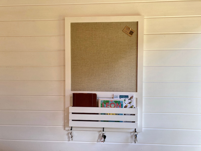 May include: White wooden wall organizer with a corkboard, a shelf for storing items, and hooks for hanging keys. The corkboard has a brown linen texture and a small brown paper note attached to it. The shelf has a brown leather notebook and a colorful postcard with the word "LEON" on it.