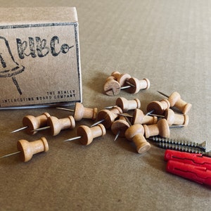 May include: A box of wooden push pins with the text "RibCo The Really Interesting Board Company" on the box. The push pins are scattered on a brown surface with two red plastic wall anchors and two screws.