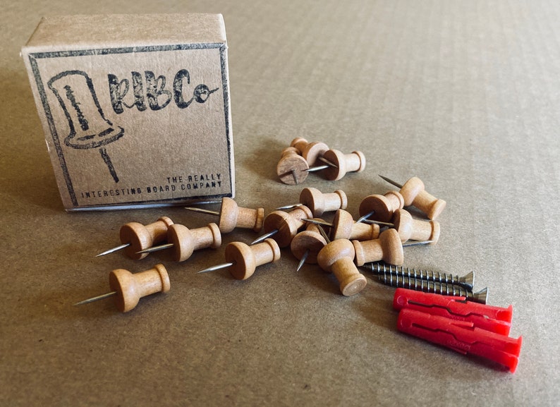 May include: A box of wooden push pins with the text "RibCo The Really Interesting Board Company" on the box. The push pins are scattered on a brown surface with two red plastic wall anchors and two screws.