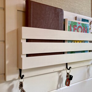 May include: White wooden wall mounted key holder with two hooks and three keys hanging from one hook. The key holder has two shelves for storing items like books or mail. The shelves are made of white wood with a natural wood grain finish. The key holder is mounted on a white wall.