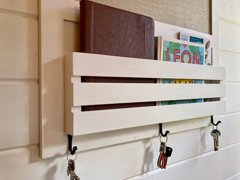 May include: White wooden wall mounted key holder with two hooks and three keys hanging from one hook. The key holder has two shelves for storing items like books or mail. The shelves are made of white wood with a natural wood grain finish. The key holder is mounted on a white wall.