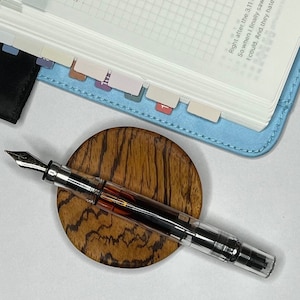 May include: A clear fountain pen with a silver nib rests on a round, wooden pen rest with a zebrawood grain pattern. A light blue planner with labeled tabs and handwritten notes is in the background.