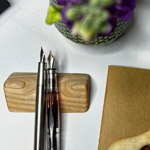 May include: Two silver fountain pens with black ink cartridges are displayed in a wooden pen holder. The pens are on a brown surface with a wooden triangle with a red circle in the center.