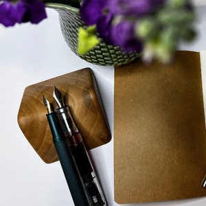 May include: A brown leather bound notebook with a blank white page open. A silver fountain pen is resting on the open page. Two other fountain pens are in a wooden pen holder.
