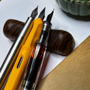 May include: Three fountain pens, one yellow, one clear with red ink, and one silver, are displayed on a wooden pen holder. The pens are resting on a brown paper surface.
