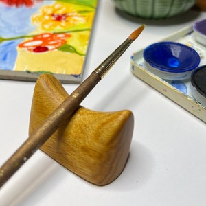 May include: A wooden paintbrush holder with a paintbrush resting on it. The holder is light brown and has a smooth, rounded surface. The paintbrush is made of natural wood and has a gold metal ferrule. The paintbrush is resting on the holder at a slight angle.