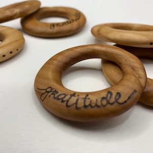 May include: Five wooden rings with the word "gratitude" inscribed on one of the rings. The rings are stacked on top of each other.