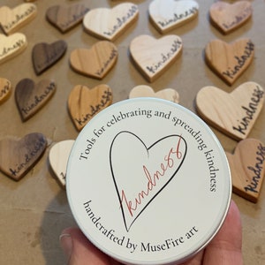 May include: A white round tin with a red heart and the word "kindness" written inside. The tin has the text "Tools for celebrating and spreading kindness handcrafted by MuseFire art" written around the heart.