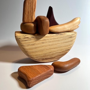 May include: A wooden sculpture composed of various shapes and colors. The base is a light-colored, half-moon shape. Several wooden pieces, including a tall, textured piece, are arranged on top. Additional pieces are scattered below.
