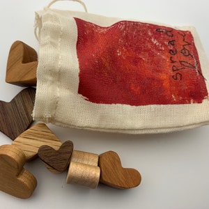 May include: A small white cloth bag with the words "Spread Love" printed on it. The bag is spilling out wooden heart-shaped ornaments in various shades of brown.
