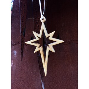 May include: A wooden star ornament with a light brown finish and a white string for hanging. The star has a pointed design with a cutout in the center.