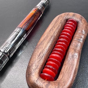May include: A clear and silver fountain pen with an amber-colored ink reservoir is positioned next to a wooden massage tool with red beads. The pen has a silver nib and a detailed design. The massage tool is made of wood and has a curved shape.