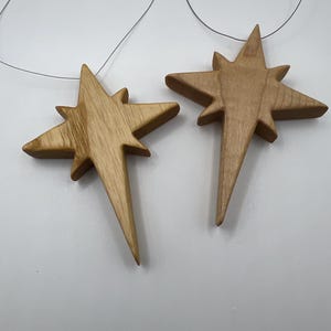 May include: Two wooden star ornaments with a natural wood grain finish. Each star has eight points and is suspended by a thin, gray string. The stars are displayed against a white background.