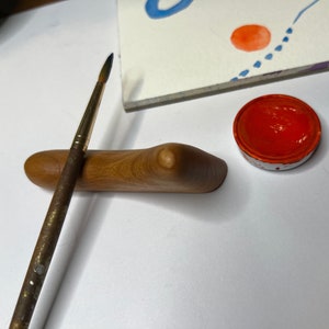 May include: A wooden paintbrush holder with a natural wood finish. A paintbrush is resting in the holder. A small red paint pot is on the table next to the holder.