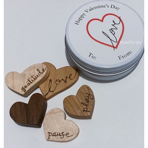 May include: A Valentine's Day gift set featuring wooden heart-shaped tokens with words like "gratitude" and "love", alongside a tin with "Happy Valentine's Day" and "love" text. The hearts are various wood tones.