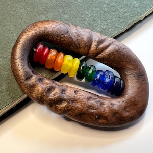 May include: A dark brown, oval-shaped wooden frame with a textured, carved surface. Inside the frame, a single horizontal string holds a series of small, translucent, circular beads arranged in a rainbow color sequence of red, orange, yellow, green, blue, and dark purple.