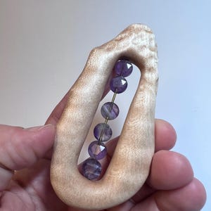 May include: A light-colored wooden sculpture with a central opening, holding a strand of faceted purple beads. The beads are strung on a thin gold wire. The sculpture has a smooth, organic shape.