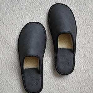 May include: A pair of dark gray leather slippers with a soft, cream-colored interior. The slippers have a rounded toe and a black trim with white stitching. A decorative bee emblem is embossed on the heel.