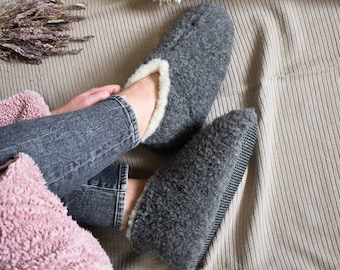 May include: A pair of gray wool slippers with a white fleece lining. The slippers are worn by a person sitting on a beige textured blanket.