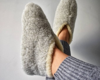 May include: A pair of gray fuzzy slippers with a white trim. The slippers are worn by a person wearing gray pants.