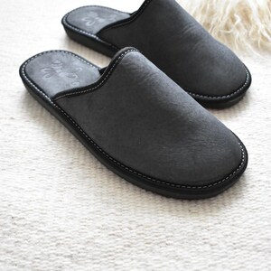 May include: A pair of charcoal gray slippers with black soles and white stitching. The slippers have a soft, textured appearance and a simple, classic design. The slippers are made of a soft material and are designed for indoor use.