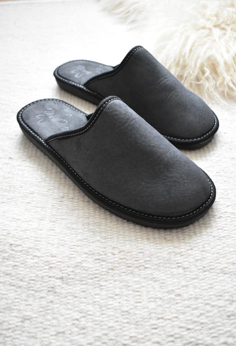 May include: A pair of charcoal gray slippers with black soles and white stitching. The slippers have a soft, textured appearance and a simple, classic design. The slippers are made of a soft material and are designed for indoor use.
