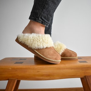 May include: Brown suede slippers with cream shearling lining and trim. The slippers have a stitched sole and are shown on a wooden bench. These indoor slippers offer warmth and comfort for the feet.