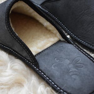 May include: Close-up of a pair of black leather slippers with a soft, cream-colored interior. The slippers have a stitched design and a bee emblem on the insole. The slippers are size 42.