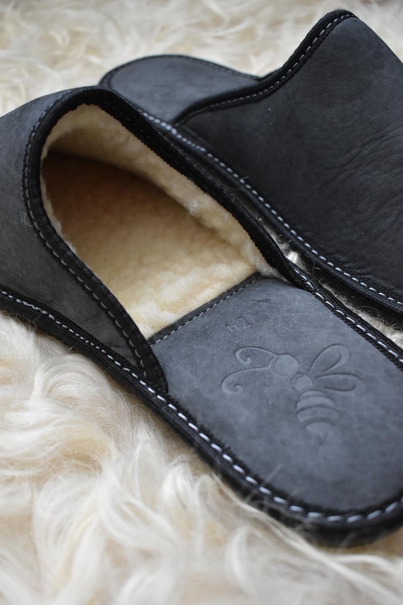 May include: Close-up of a pair of black leather slippers with a soft, cream-colored interior. The slippers have a stitched design and a bee emblem on the insole. The slippers are size 42.